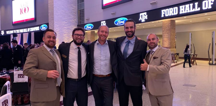 Five men dressed in suits stand together inside a large hall with "Ford Hall of Champions" and Texas A&M signage visible behind them. Four smile at the camera while one gives a thumbs-up, representing Patriot Pool and Spa at the event.