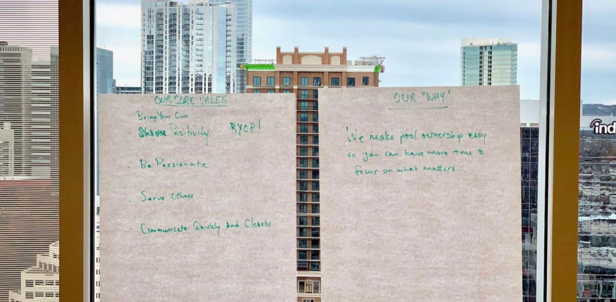 Two large sheets of paper are taped to a window overlooking city buildings. Each page features handwritten notes in green marker outlining Patriot Pool and Spa’s core values and company mission. The cityscape is visible through the glass.