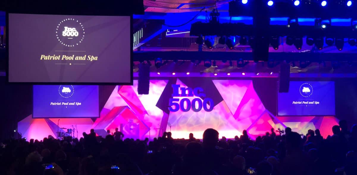 A stage displays "Inc. 5000" in large letters, with screens highlighting Patriot Pool and Spa. The audience is seated in a dimly lit venue, illuminated by purple and blue lighting.