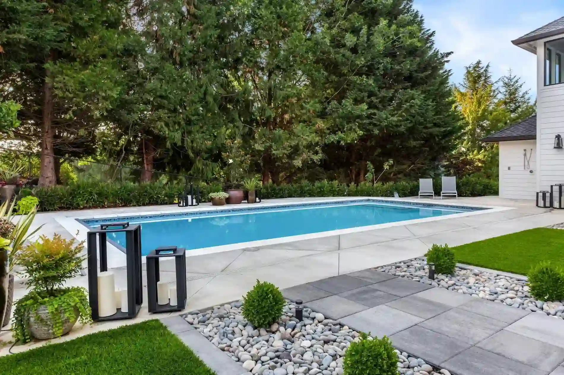 A modern backyard by Patriot Pool and Spa features a rectangular swimming pool, stone paving, and lush greenery. Decorative lanterns, small shrubs, and lounge chairs line the poolside, framed by tall trees in the background.