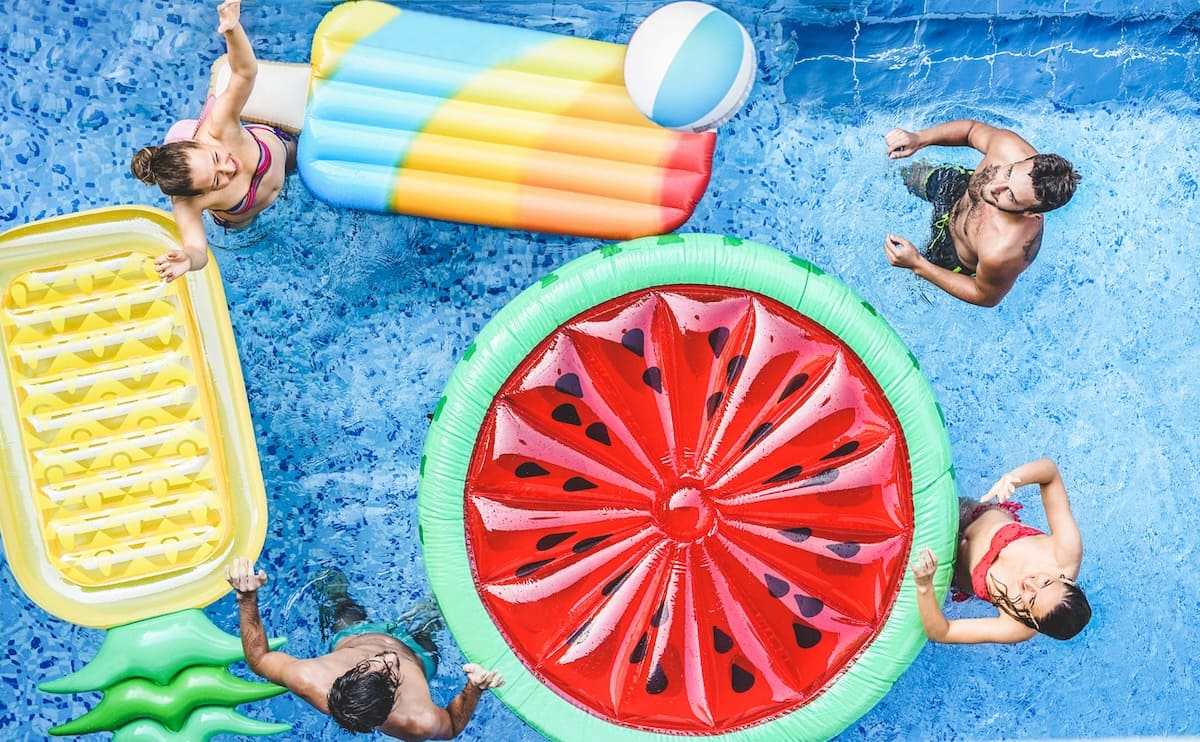 Four people are in a sparkling Patriot Pool and Spa swimming pool with three large inflatable floats shaped like a popsicle, a slice of watermelon, and pineapple. One person holds a beach ball while the clear blue water glistens around them.