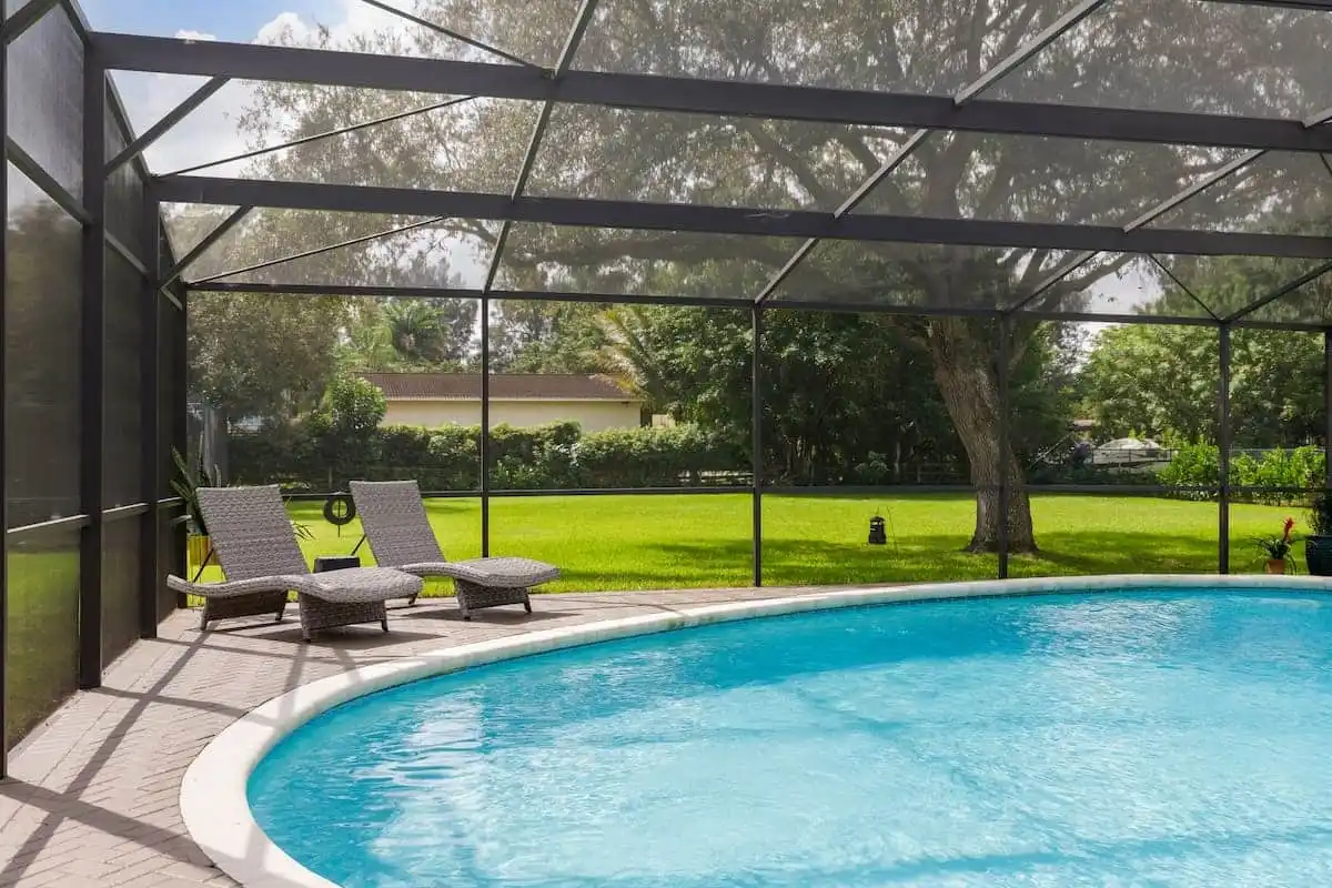A screened-in outdoor pool from Patriot Pool and Spa features clear blue water and a paved deck. Two wicker lounge chairs invite relaxation, with green grass and trees visible just beyond the enclosure.