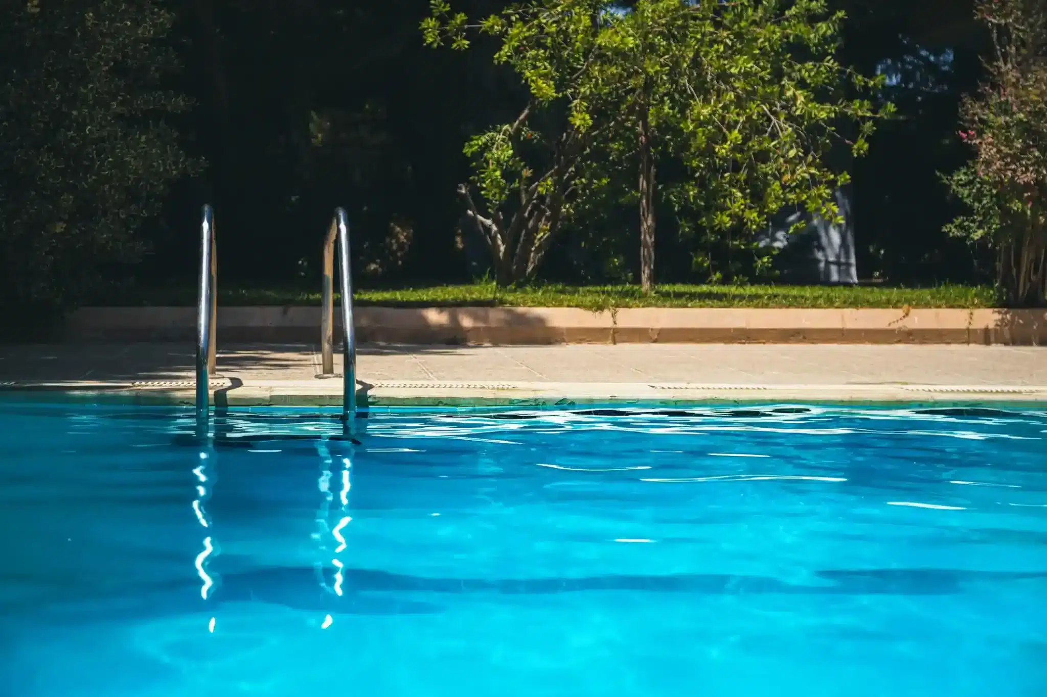A swimming pool with clear blue water, a metal ladder on the left side, and a concrete deck. Green grass, trees, and shrubs are visible in the background under bright sunlight—another serene scene by Patriot Pool and Spa.