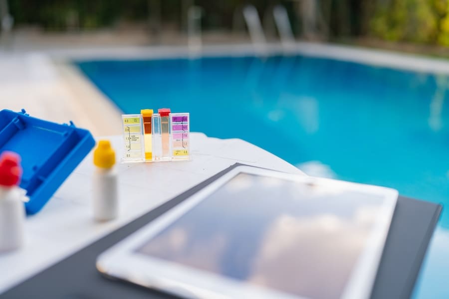 A close-up of Patriot Pool and Spa water testing equipment, color comparison kits, and a tablet on a poolside table, with a clear blue swimming pool in the background.
