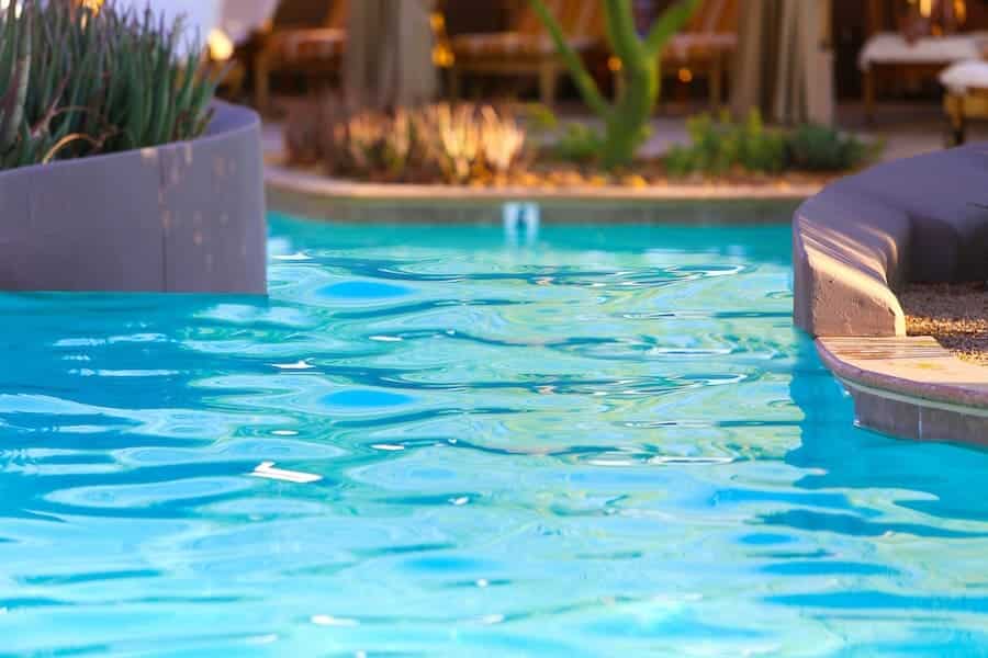 Clear blue water in a swimming pool with curved edges by Patriot Pool and Spa sits beside landscaped plants. In the background, blurred lounge chairs and greenery suggest a tranquil outdoor leisure area.