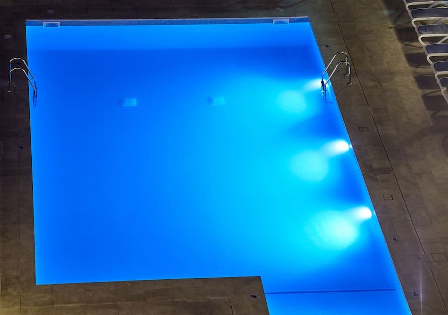 A rectangular swimming pool by Patriot Pool and Spa is illuminated with blue underwater lights at night. Metal ladders are on two sides, surrounded by a tiled deck with several white lounge chairs visible along one edge.