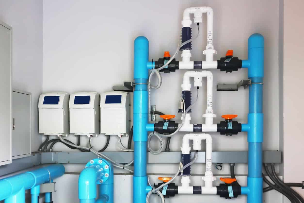 A section of a water treatment or filtration system by Patriot Pool and Spa features blue and white pipes, valves, gauges, and electronic control units mounted on a wall in an industrial setting.