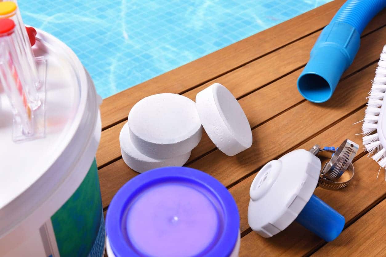 Pool cleaning supplies from Patriot Pool and Spa rest on a wooden deck by the pool, including chlorine tablets, a blue and white floating dispenser, a brush, hose, and large container.