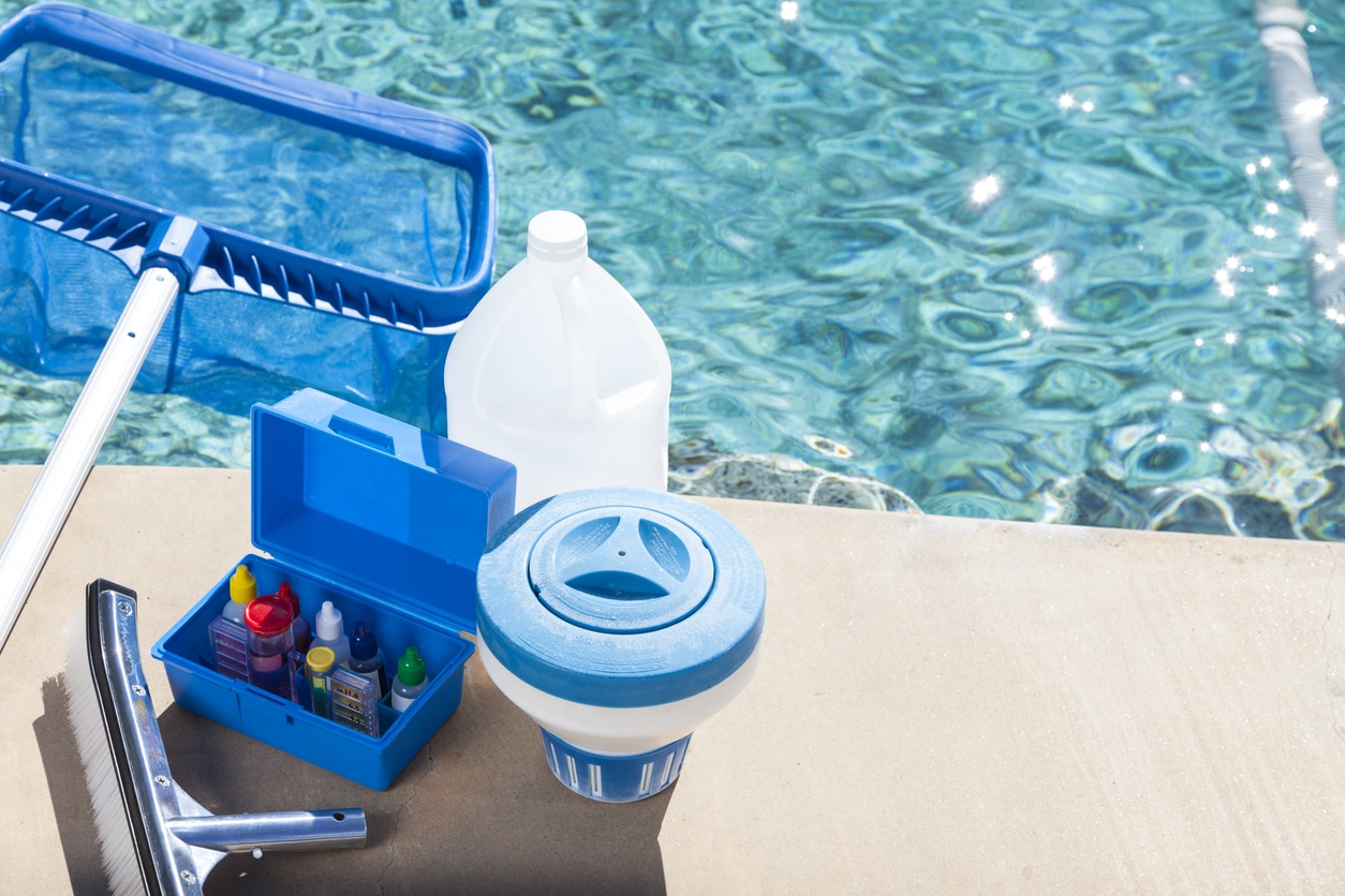 Patriot Pool and Spa cleaning supplies, including a net, liquid bottle, test kit with colored bottles, chlorine dispenser, and brush, are arranged on a pool deck next to clear blue pool water.