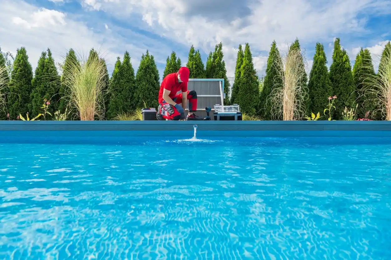A person wearing red clothing kneels next to an outdoor swimming pool, inspecting the water. The pool, serviced by Patriot Pool and Spa, is surrounded by greenery, tall trees, and ornamental grasses under a partly cloudy sky.