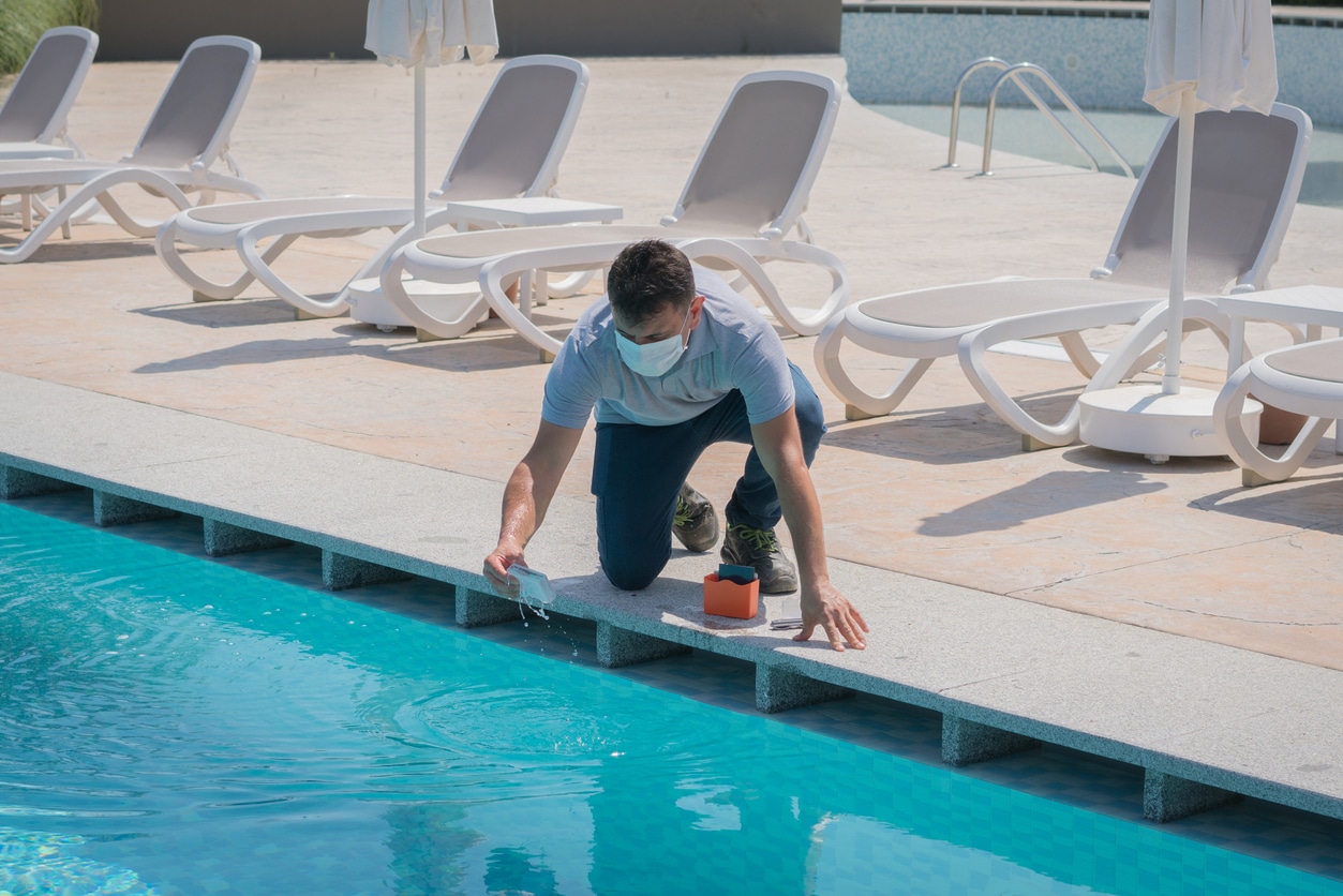 A person wearing a face mask kneels by the edge of an outdoor pool, pouring a substance into the water. Several empty lounge chairs and umbrellas are visible in the background at Patriot Pool and Spa.