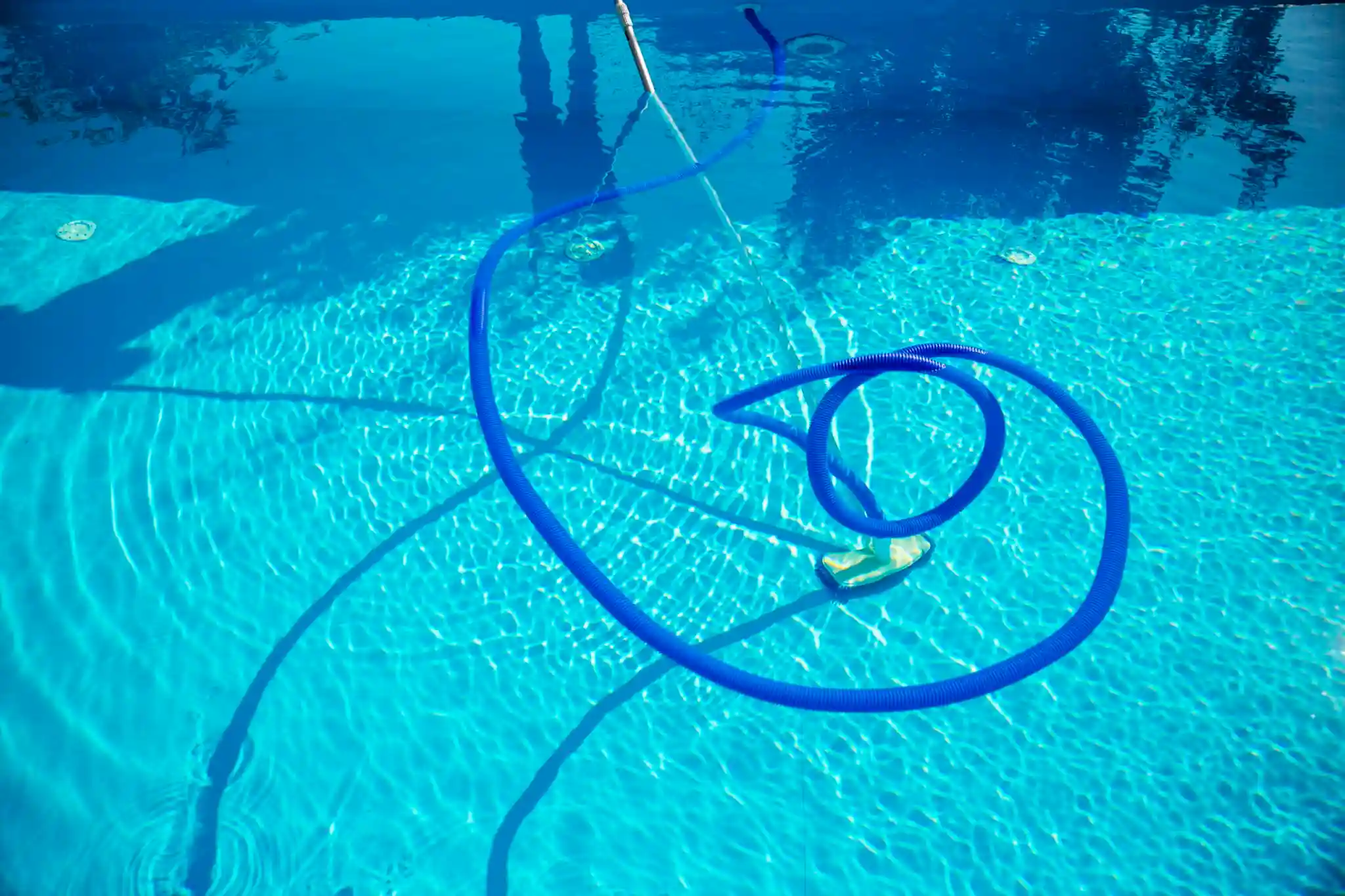 A blue pool vacuum hose coils on the surface and floor of a clear, sunlit swimming pool at Patriot Pool and Spa, creating ripples and shadows in the water.