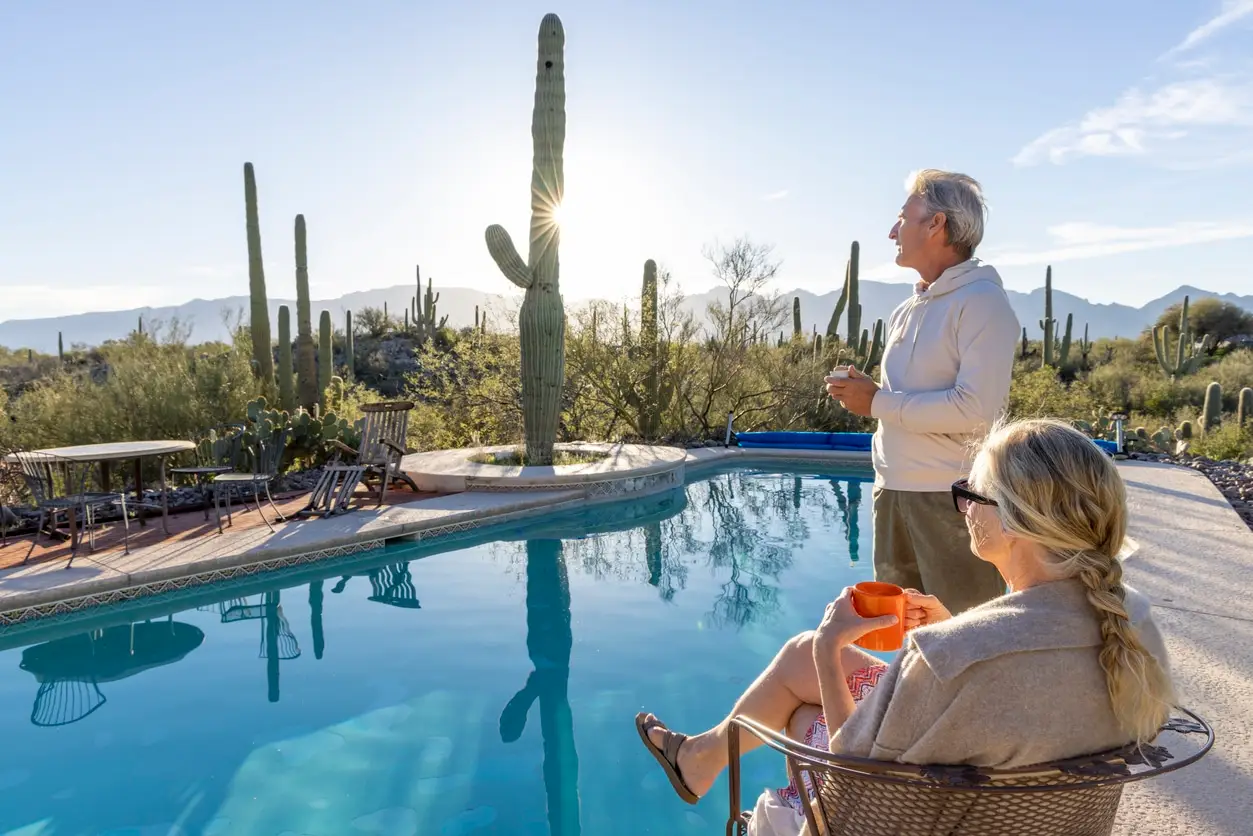 An older man and woman relax by a swimming pool in a desert setting with large cacti, enjoying the serene atmosphere. The inviting scene showcases a Patriot Pool and Spa, perfect for unwinding as the sun sets behind the cactus-dotted horizon.