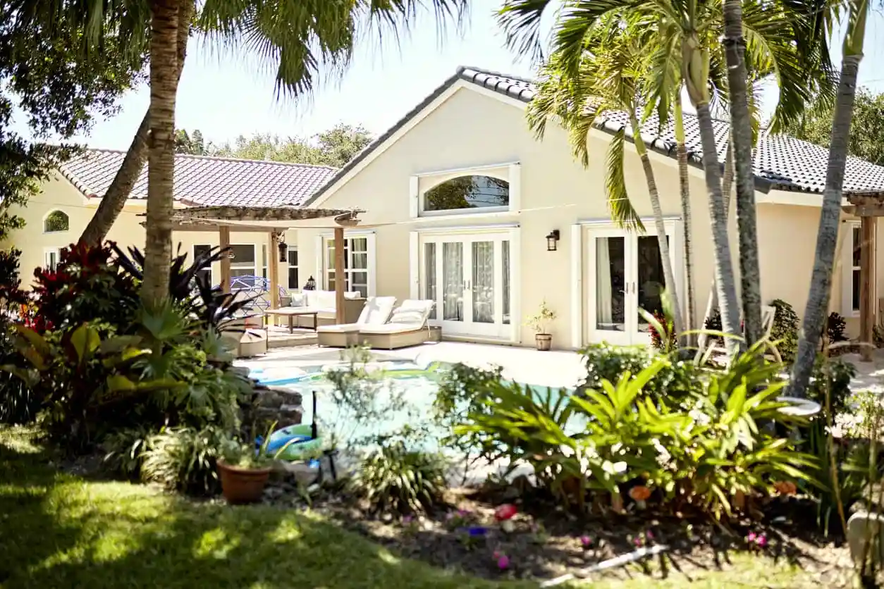 A single-story house with light-colored walls and large windows is surrounded by tropical plants. The backyard features a Patriot Pool and Spa, as well as a cozy patio area with outdoor seating.