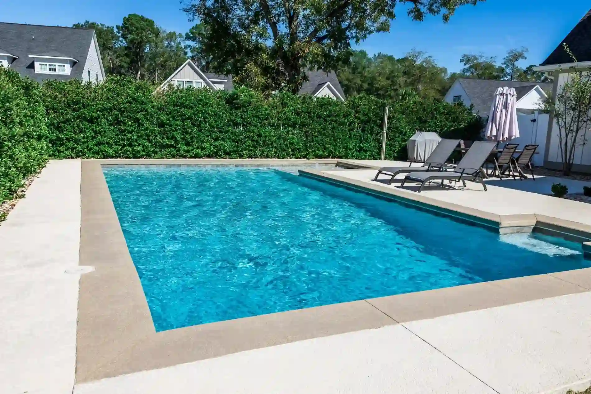 A rectangular outdoor swimming pool with clear blue water, maintained by Patriot Pool and Spa, is surrounded by a concrete deck. Several lounge chairs and an umbrella line the deck, with green hedges and houses in the background.
