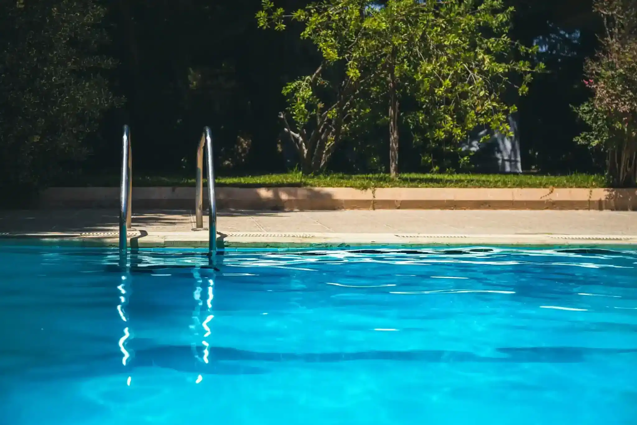 A swimming pool with clear blue water, a metal handrail ladder, and trees with green foliage in the background on a sunny day—perfectly maintained by Patriot Pool and Spa.