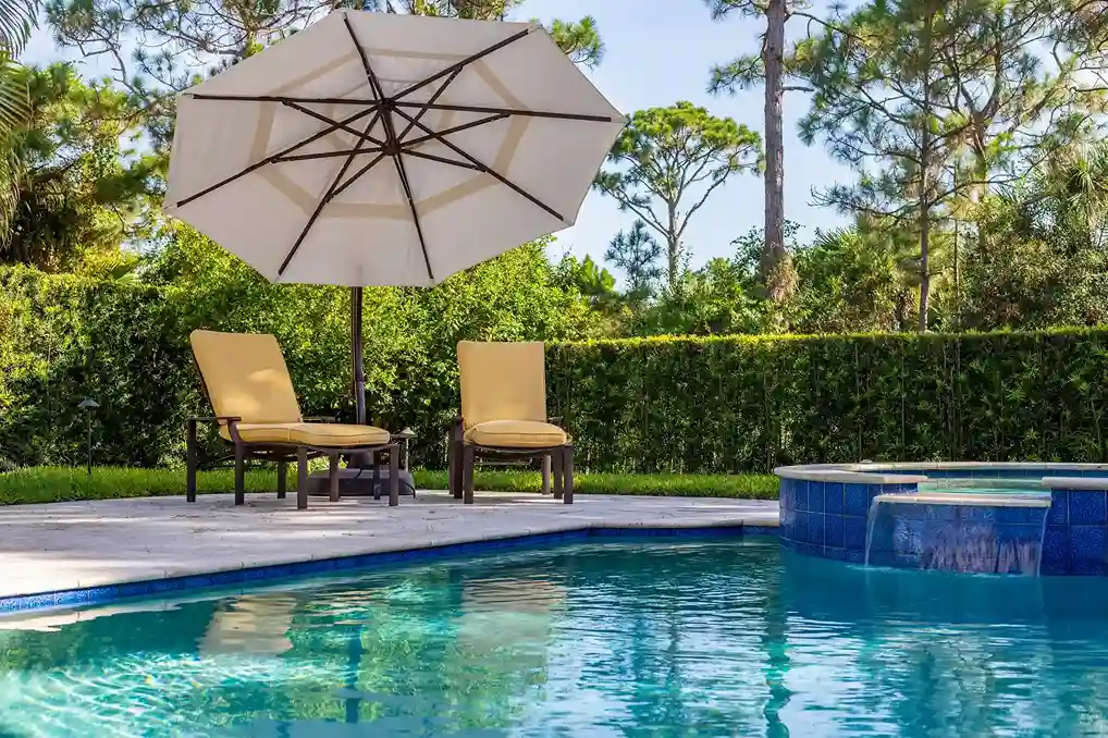 Two yellow cushioned lounge chairs sit under a large white patio umbrella beside a sparkling Patriot Pool and Spa, featuring a small attached spa and surrounded by lush greenery and trees.