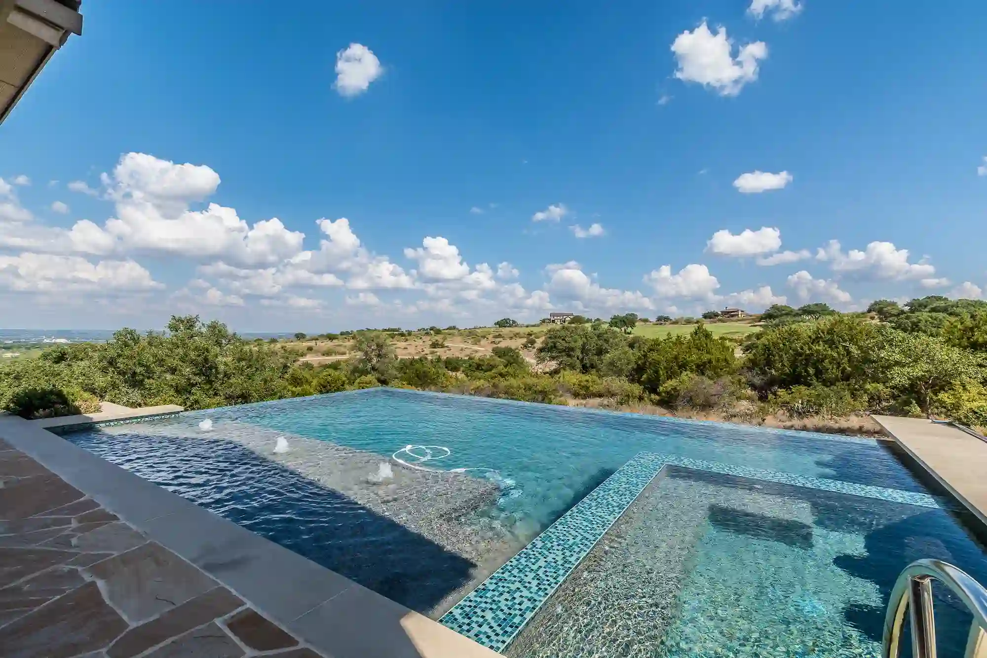 Infinity-edge swimming pool by Patriot Pool and Spa with clear blue water overlooks a hilly, green landscape under a mostly sunny sky with scattered clouds. A stone pool deck is visible in the foreground.
