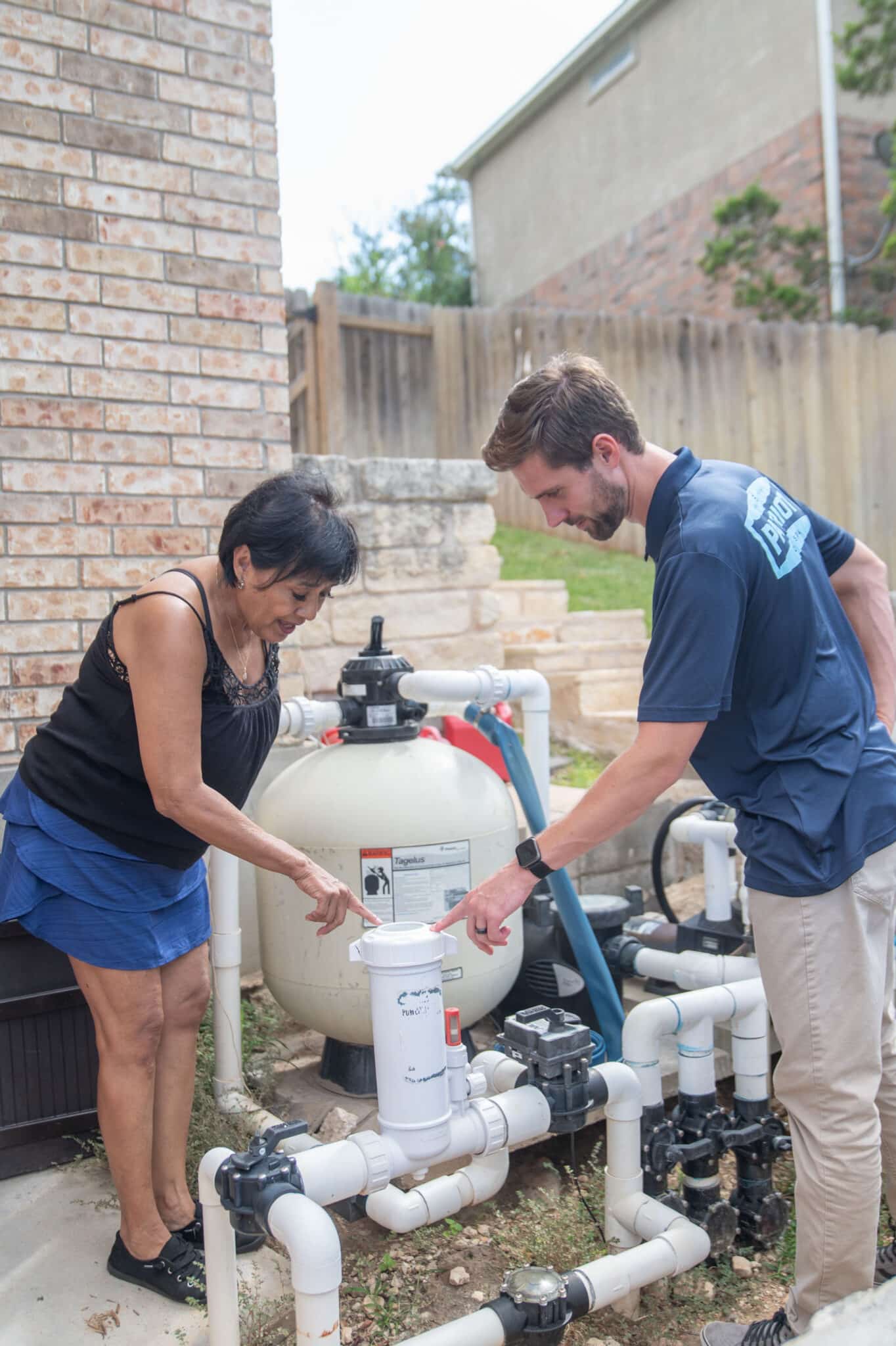 A woman and a man stand outdoors next to a pool filtration system, both pointing at a white PVC pipe among several pipes and valves—discussing equipment care for optimal pool protection. The man wears a blue uniform shirt with a logo on the back.
