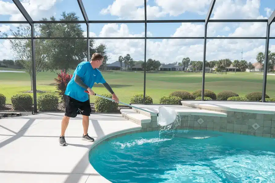 A man in a blue shirt and black shorts uses a net to clean leaves from a screened-in outdoor swimming pool on a sunny day, with Patriot Pool and Spa service ensuring the area is pristine and a golf course visible in the background.