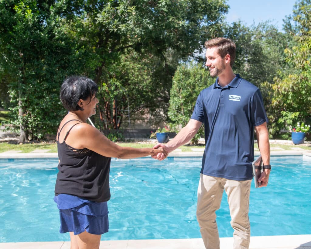 A woman in a black tank top and blue shorts shakes hands with a man in a navy polo shirt by a clean swimming pool, with trees and plants in the background on a sunny day.